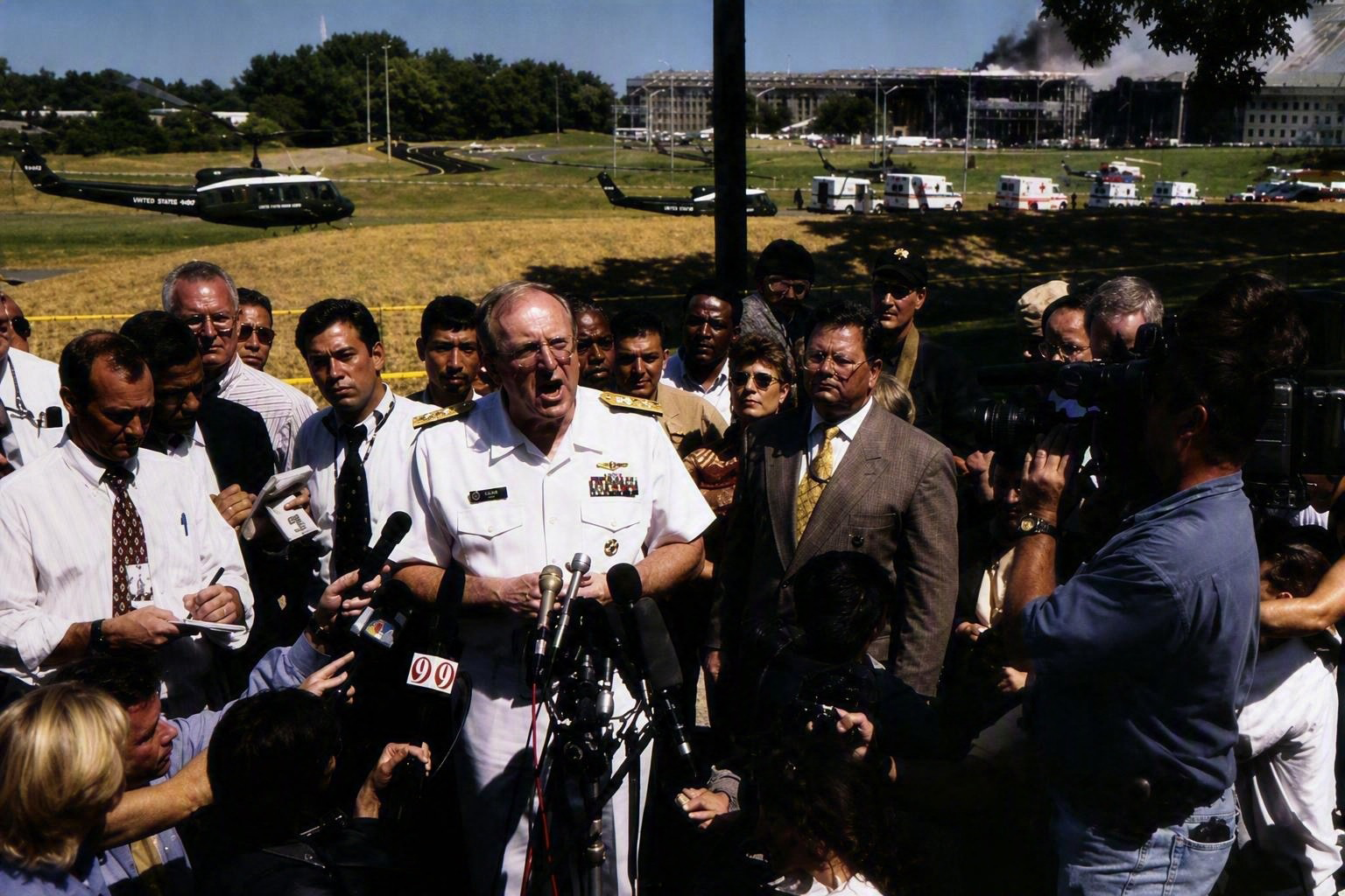 A rear admiral in service dress white speaking into a bank of microphones on a lawn, with smoke rising from the Pentagon behind him.