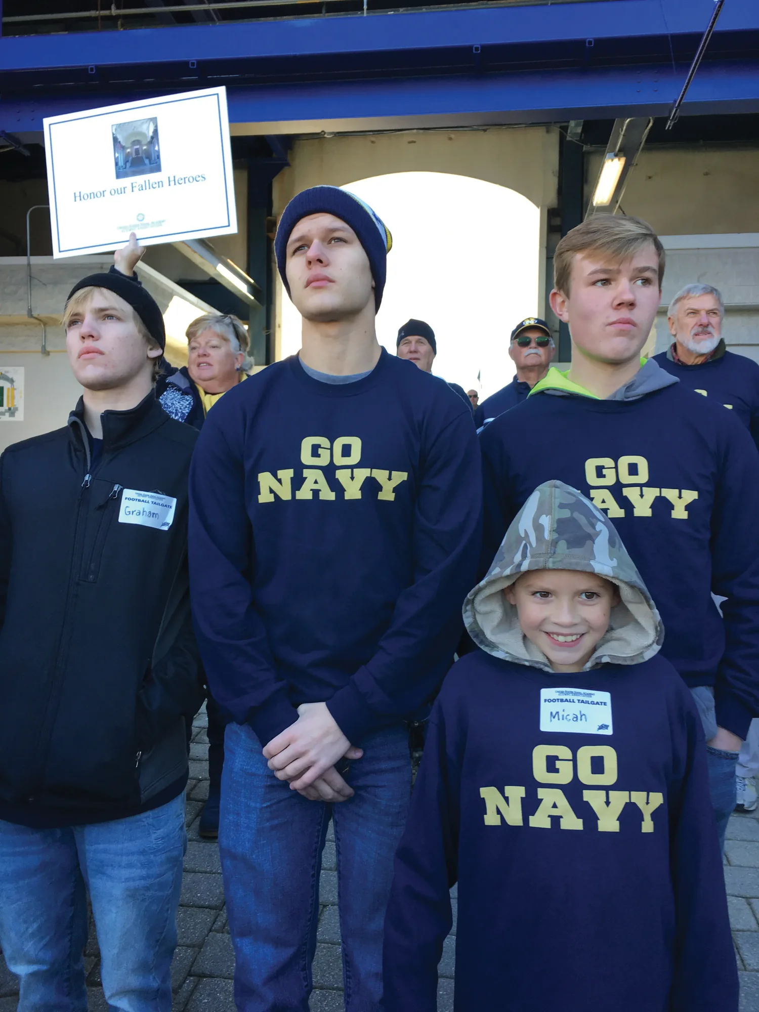 Four young members of the Michaud family in Go Navy shirts at a football tailgate, an Honor our Fallen Heroes placard held up behind them.