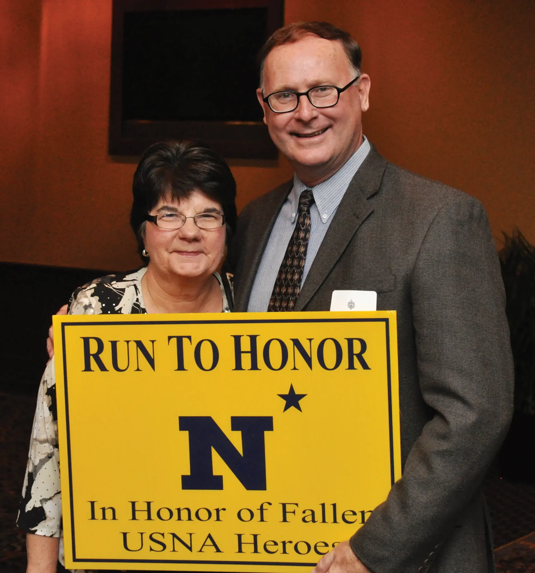 A couple holding a yellow Run to Honor placard at an indoor ceremony.